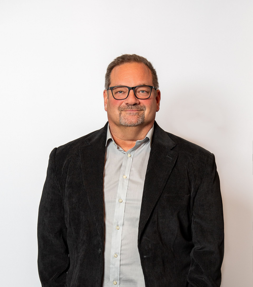 Man in glasses wearing a black jacket and light shirt, standing against a plain white background
