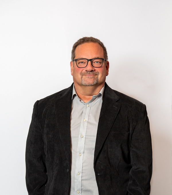 Man in glasses wearing a black jacket and light shirt, standing against a plain white background