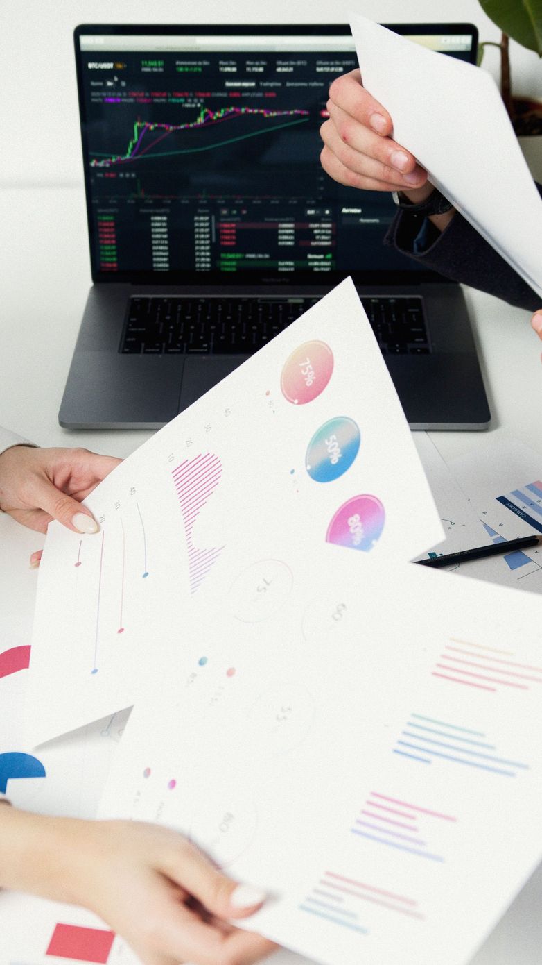 Hands reviewing printed charts and a laptop with stock market graphs on screen