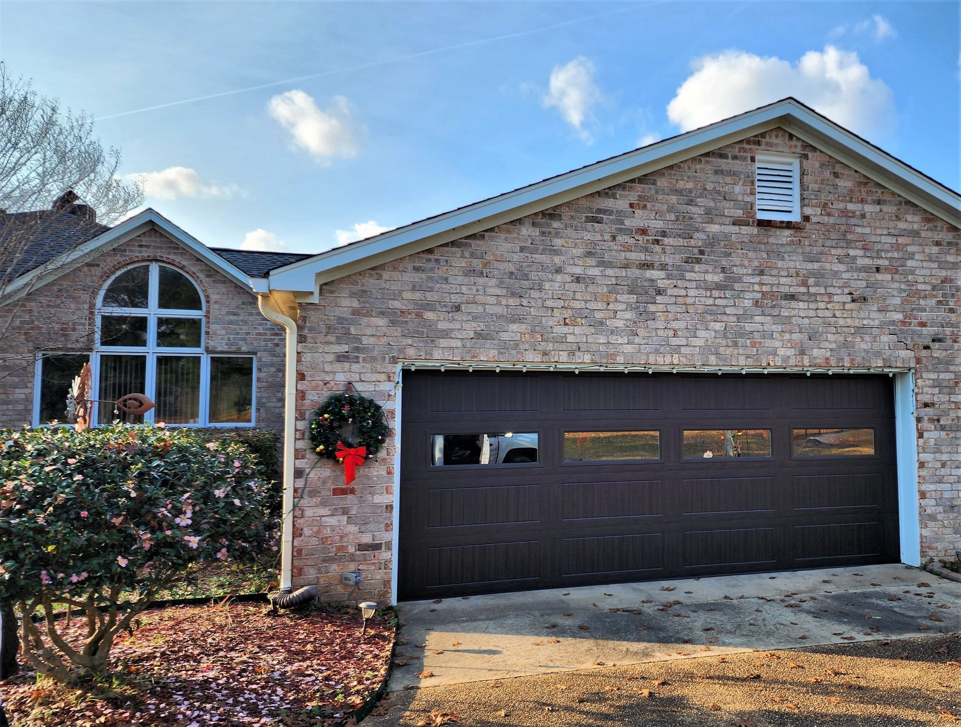 Brick house with dark brown garage door, wreath, and arched window. Sunny day.