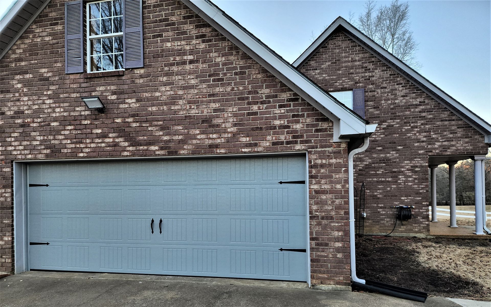 Brick house with gray garage door, window with shutters, and porch.