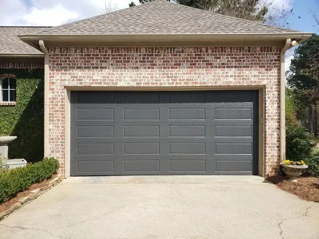 A dark gray, four-panel residential garage door set into a brick exterior wall above a concrete driveway. A dark gray, four-panel residential garage door set into a brick exterior wall above a concrete driveway.