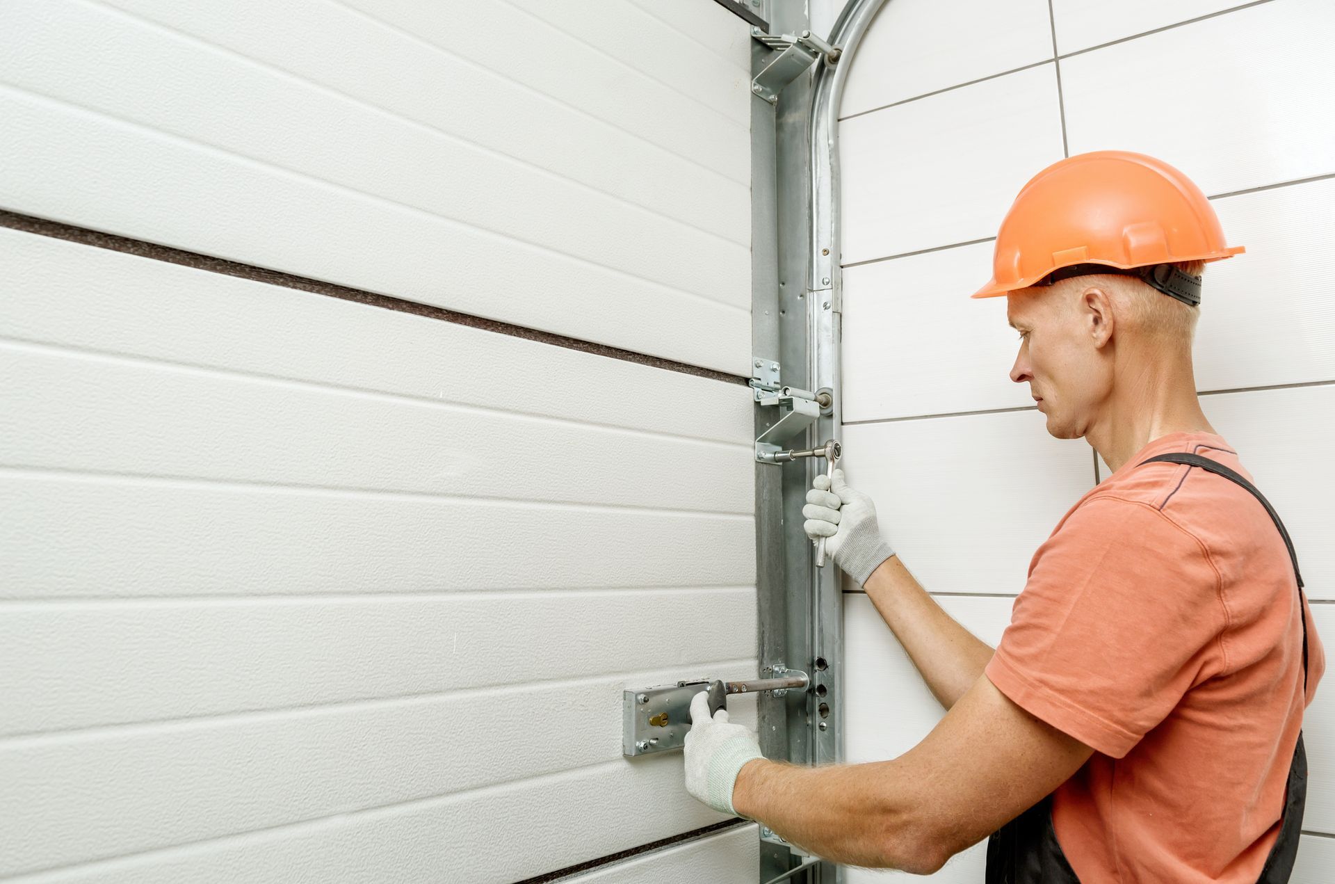 Construction worker installing a garage door. White door, orange hard hat, gloves, indoor setting.