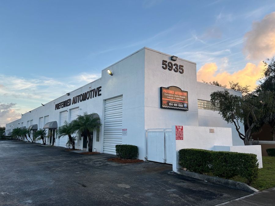 Exterior view of Preferred Automotive building, white with black signage and a sign on the right side; sky in the background.