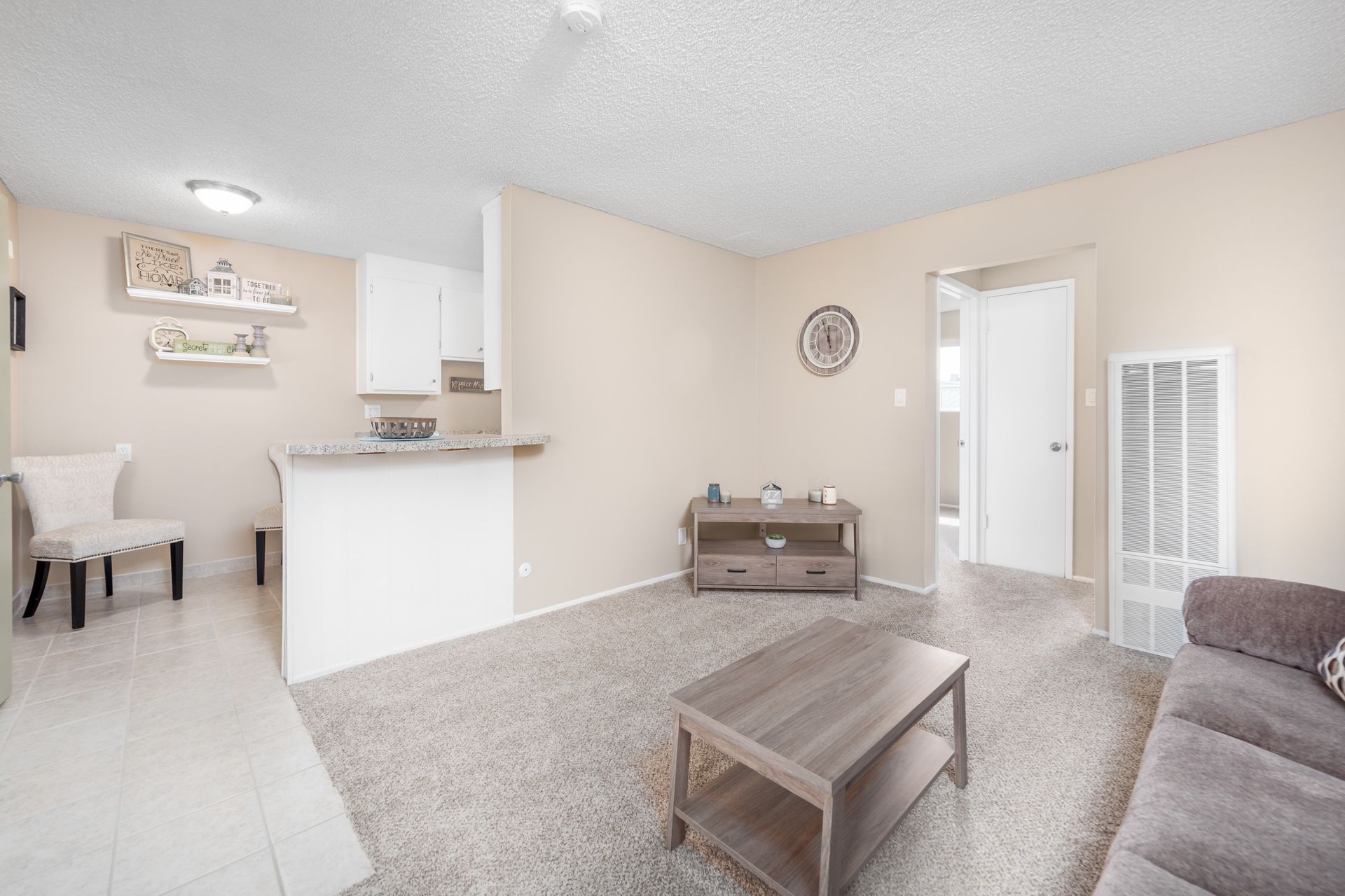 Bright beige living room with gray sofa, coffee table, carpet, and open kitchen area.