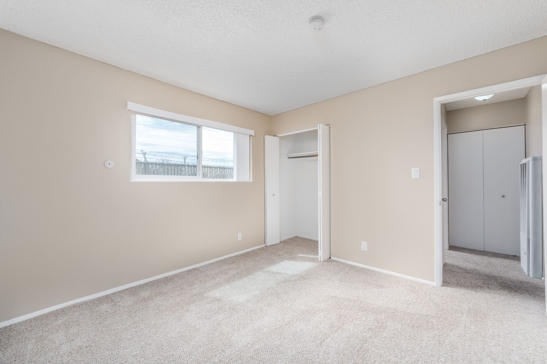 Empty beige bedroom with carpet, window, open closet, and doorway to a hall