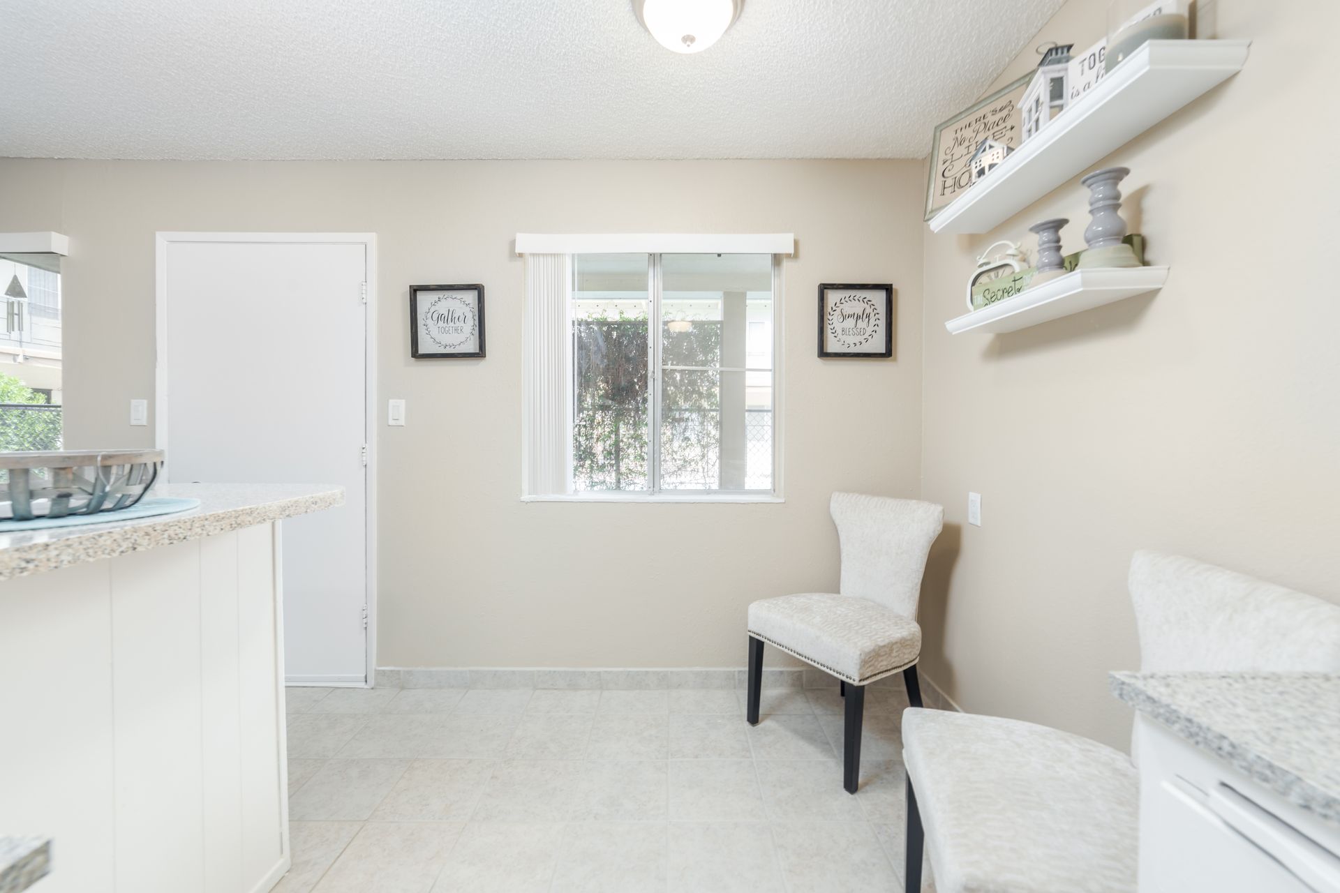 Bright bathroom with white vanity, sink, window, wall shelves, and two white chairs.