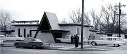 A mid-century modern funeral home with a prominent A-frame entrance and several vintage cars parked in front.