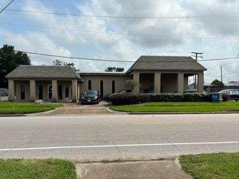 A single-story, beige commercial building with covered columned porches, fronted by a parking lot and grassy lawn.