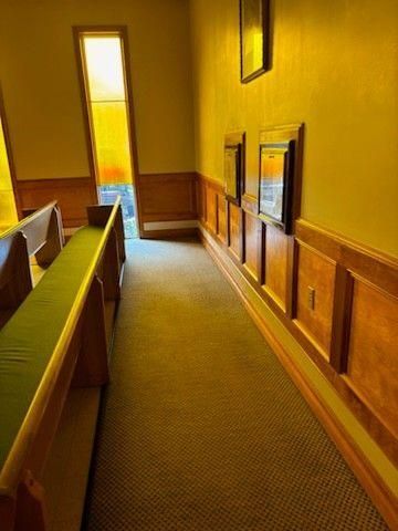 A narrow carpeted aisle in a room with wood-paneled walls, a green-cushioned pew, and tall, sunlit windows.