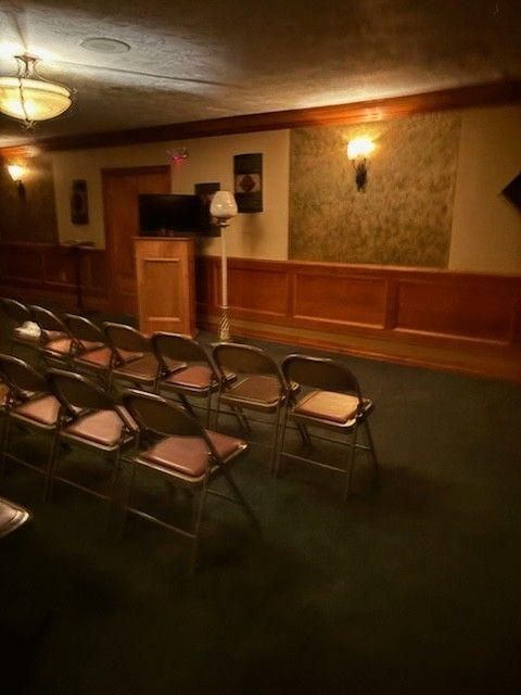 A rows of folding chairs in a dimly lit, wood-paneled room facing a small lectern with a television and lamp.