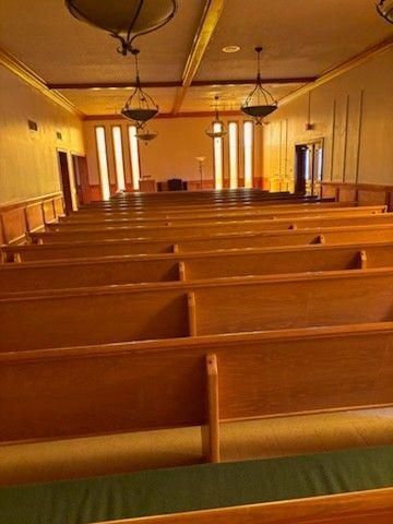 Interior of a church sanctuary with rows of wooden pews facing a raised platform illuminated by tall, vertical windows.