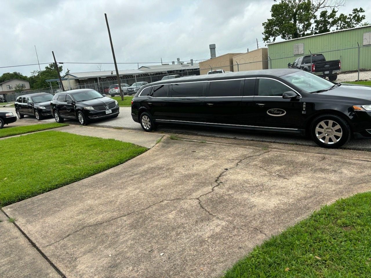 A long black limousine parked in a residential driveway, with two black SUVs parked behind it on a cloudy day.