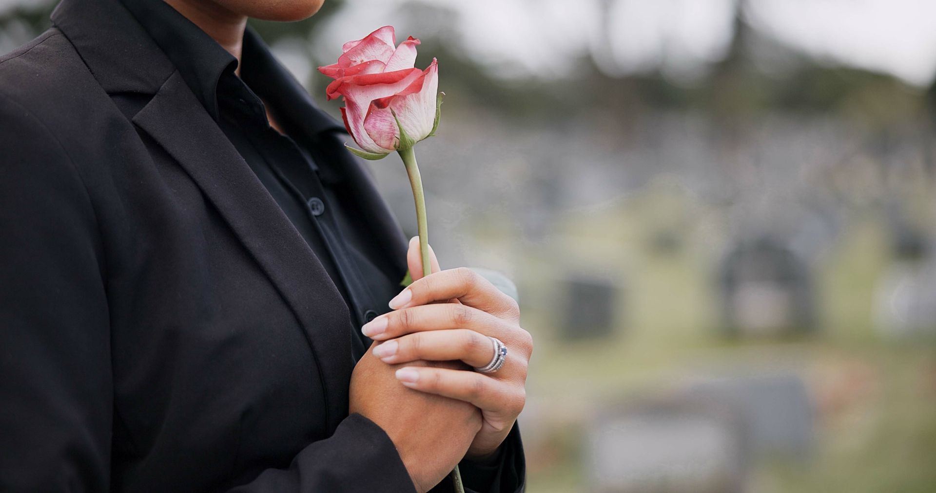 A person in a black suit stands in a cemetery, holding a single pink and white rose against their chest.