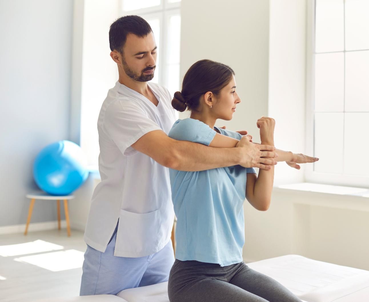 Physical therapist guiding a seated patient through arm stretches in a bright clinic room