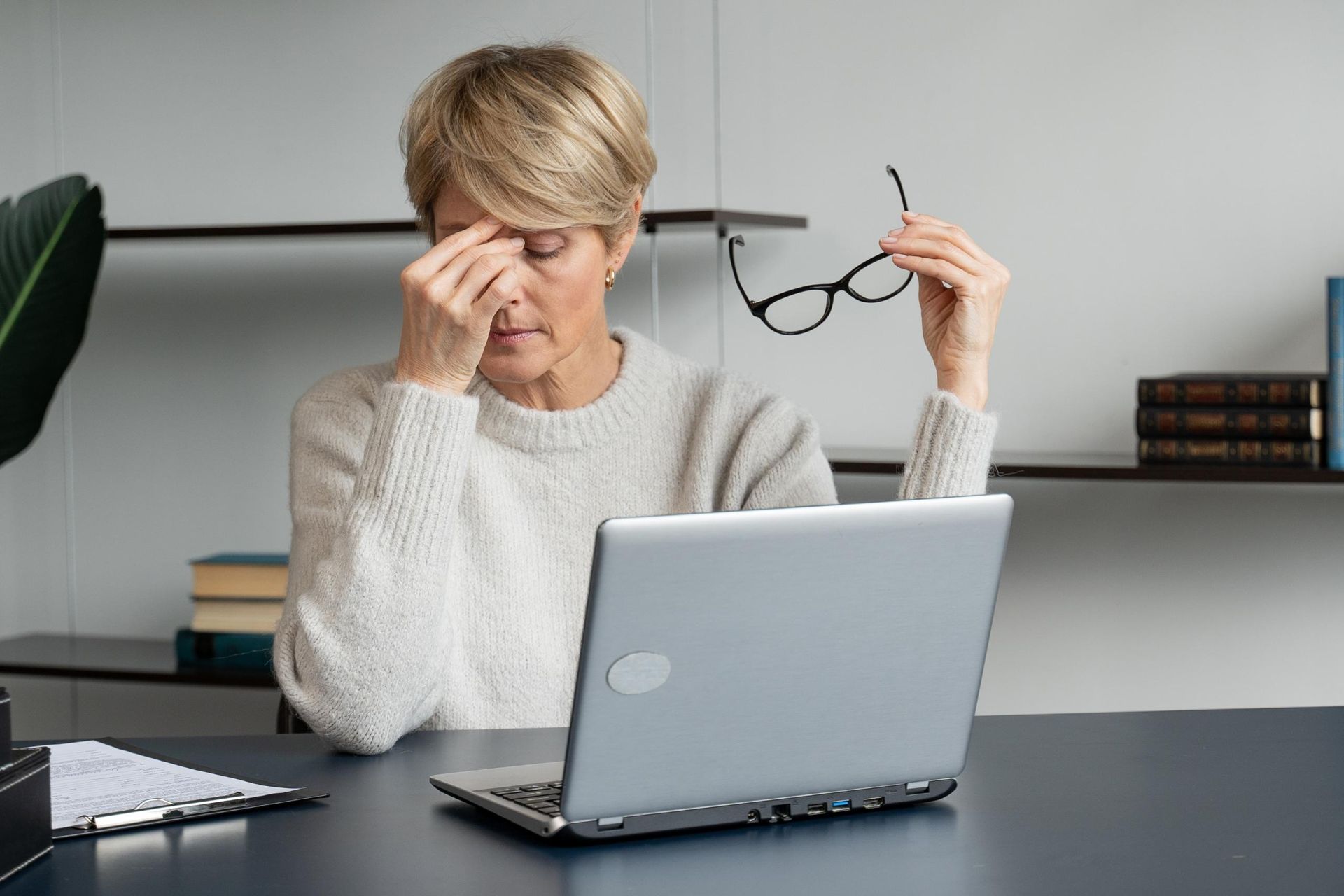Woman at a desk rubbing her eyes while holding glasses beside an open laptop