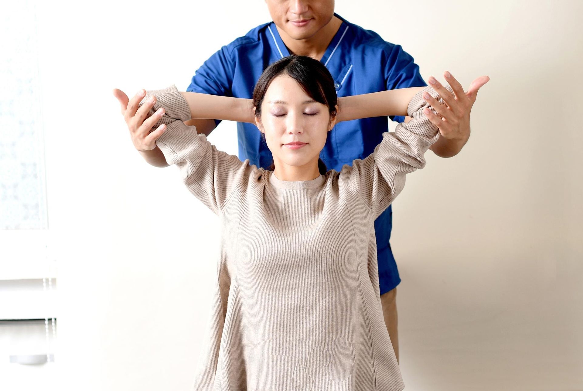 Person stretching arms forward while another stands behind assisting in a bright room