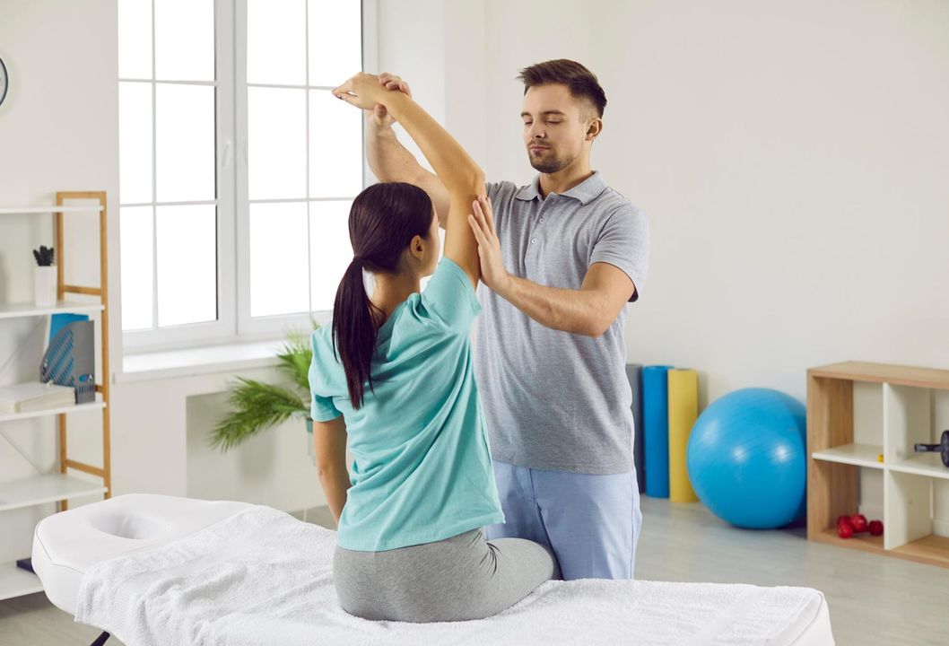 Physical therapist assisting a patient with arm exercises on a treatment table in a bright clinic room