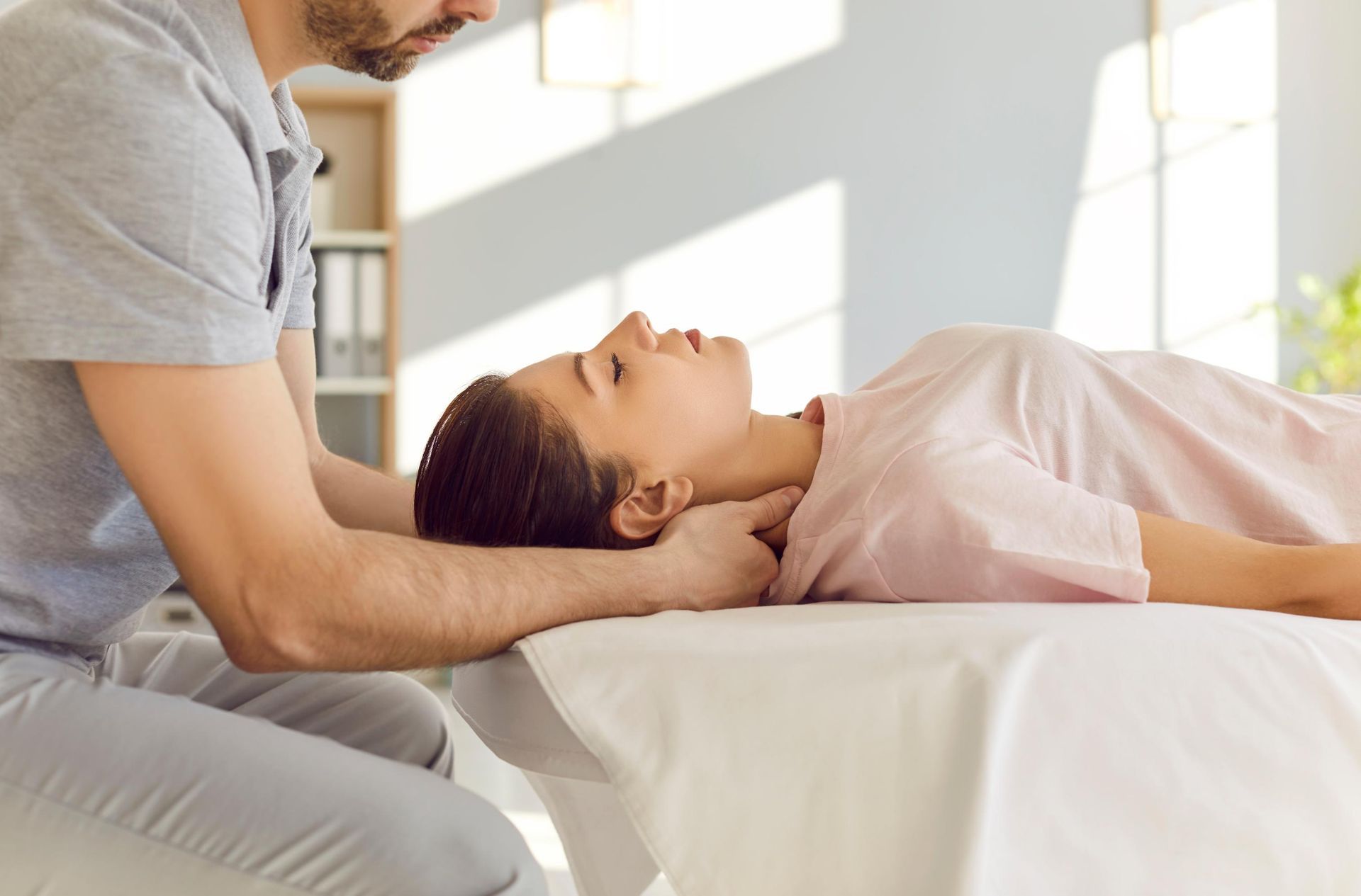 Therapist gently supports a reclining patient’s neck during a treatment session in a bright room