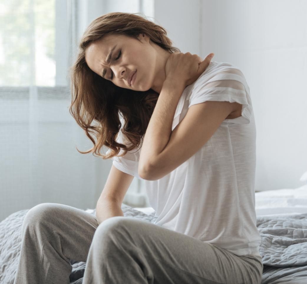 Person sitting on a bed, clutching their neck in a bright bedroom, looking uncomfortable