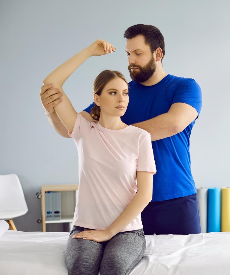 Physical therapist guiding a seated woman through a shoulder stretch in a bright clinic room
