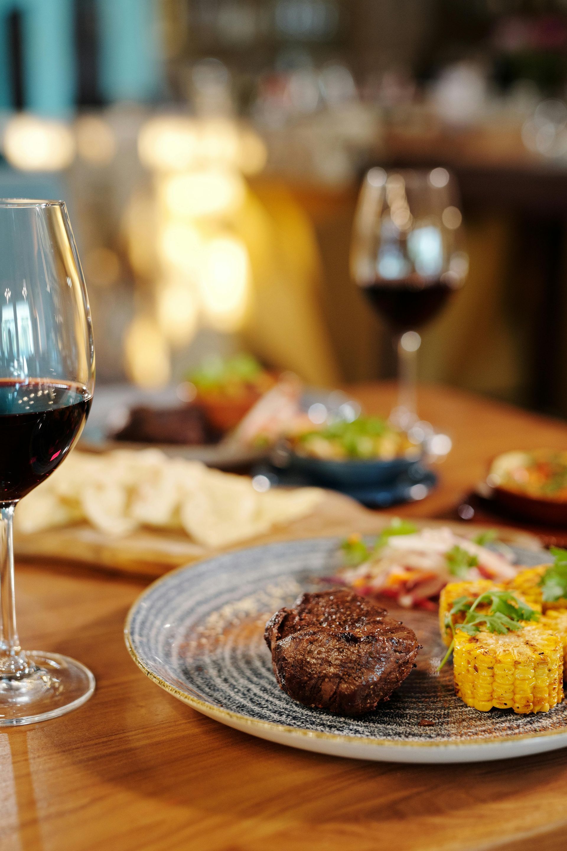 Steak dinner with grilled corn and wine glasses on a wooden table.