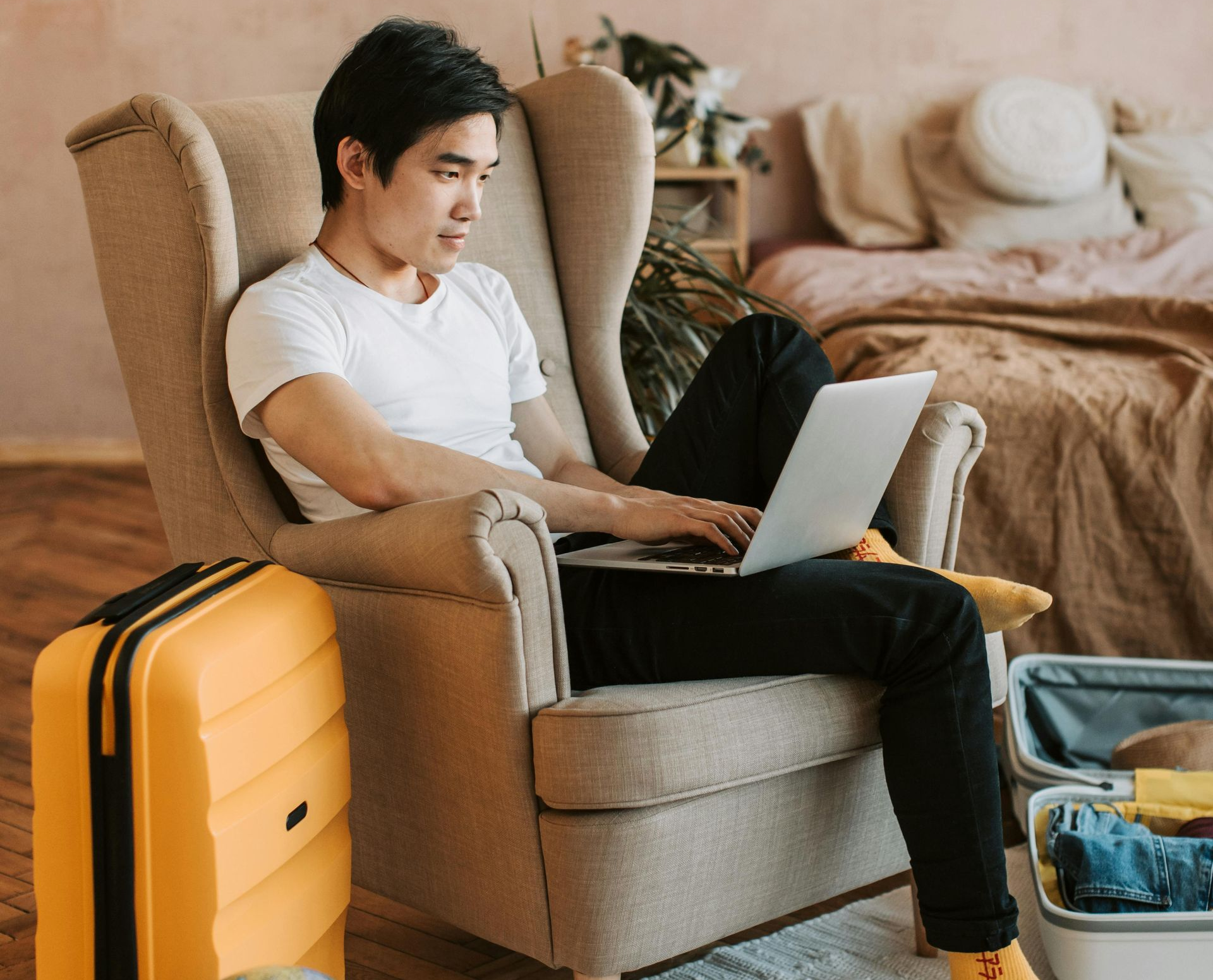 Man sitting in armchair, working on laptop; suitcase and open luggage nearby in a bedroom.