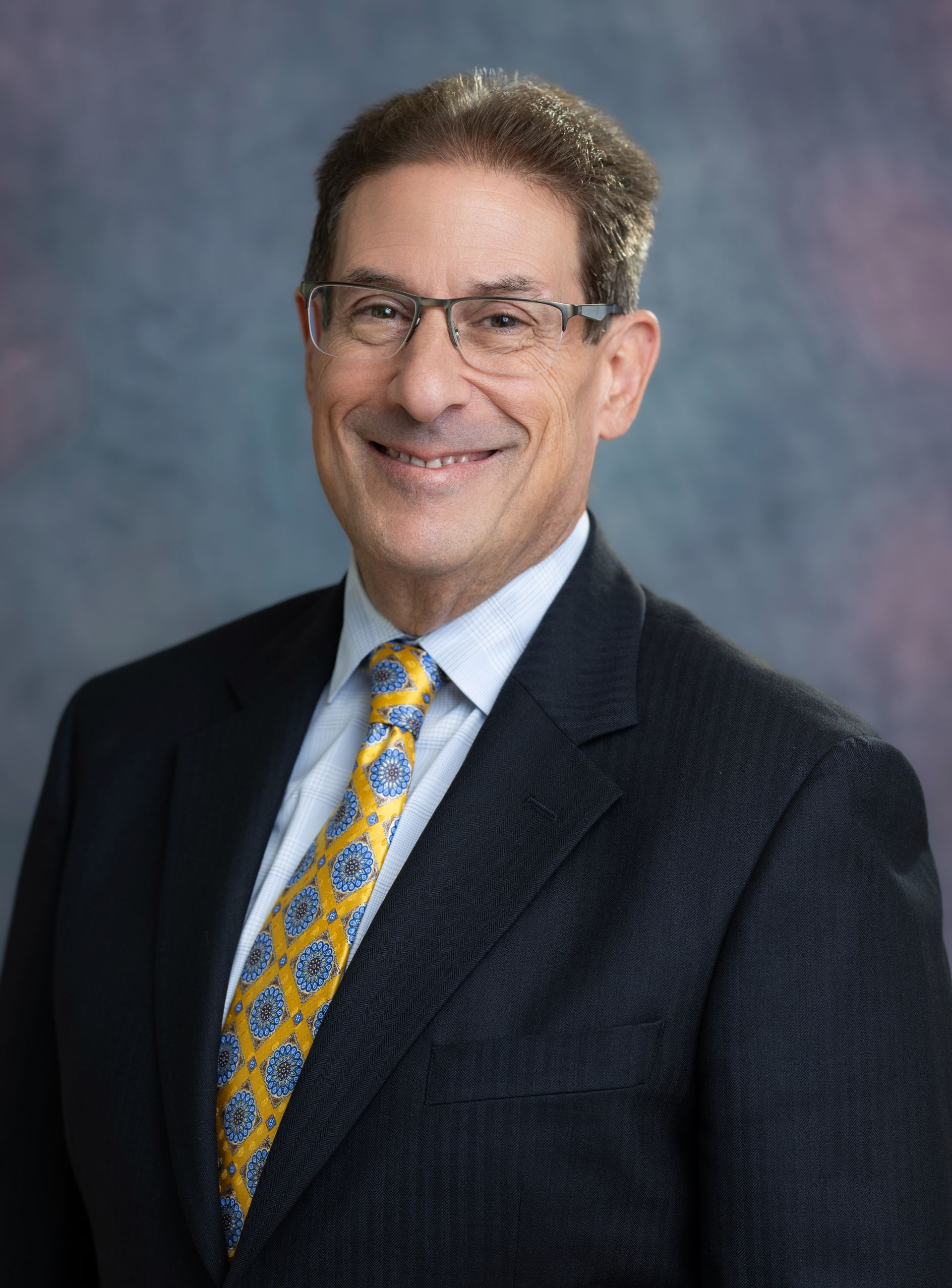Smiling man in a suit and tie, wearing glasses. He has light skin, brown hair, and a dark red tie.