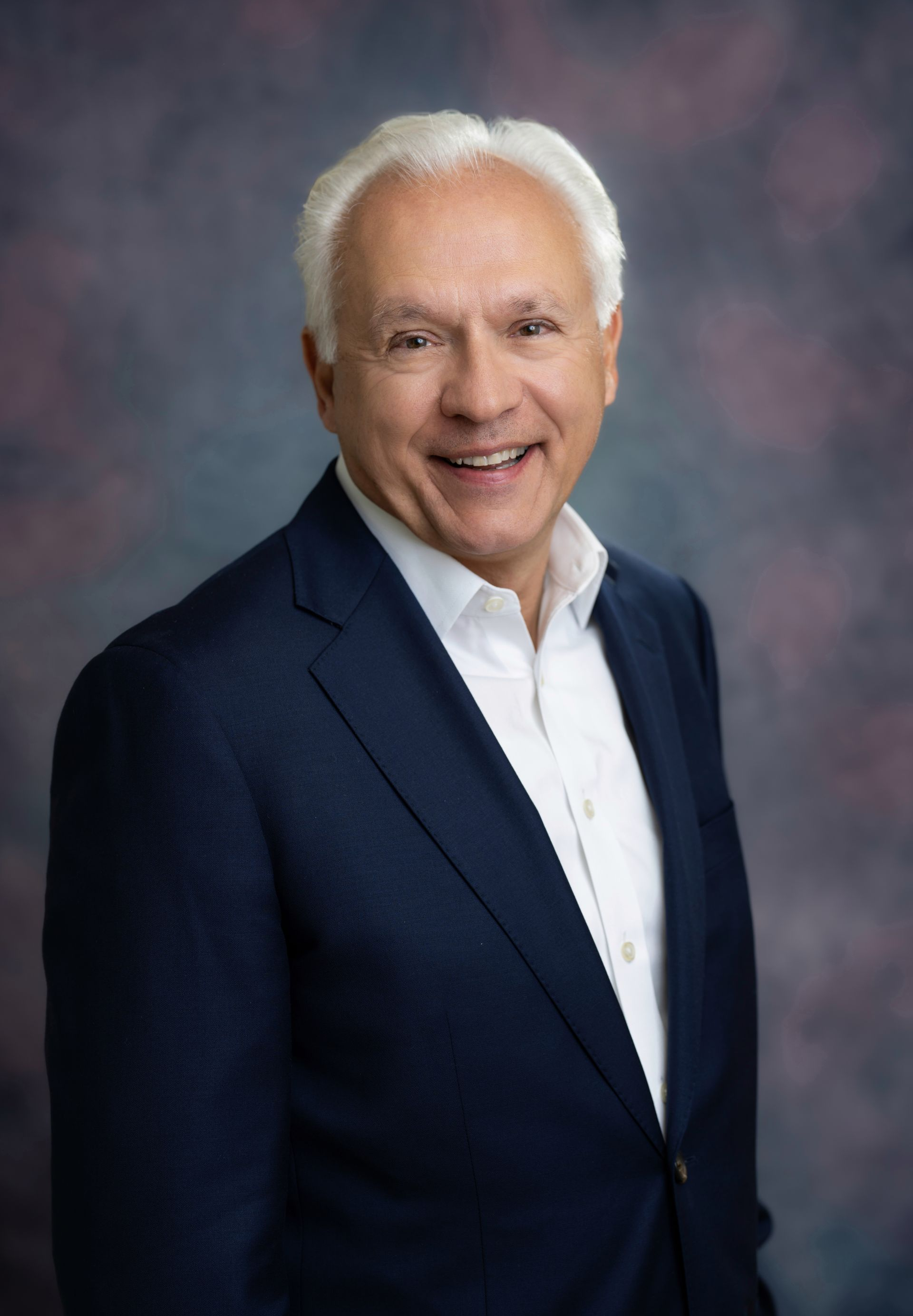 Smiling older man in a navy suit and tie, white hair, against a dark background.