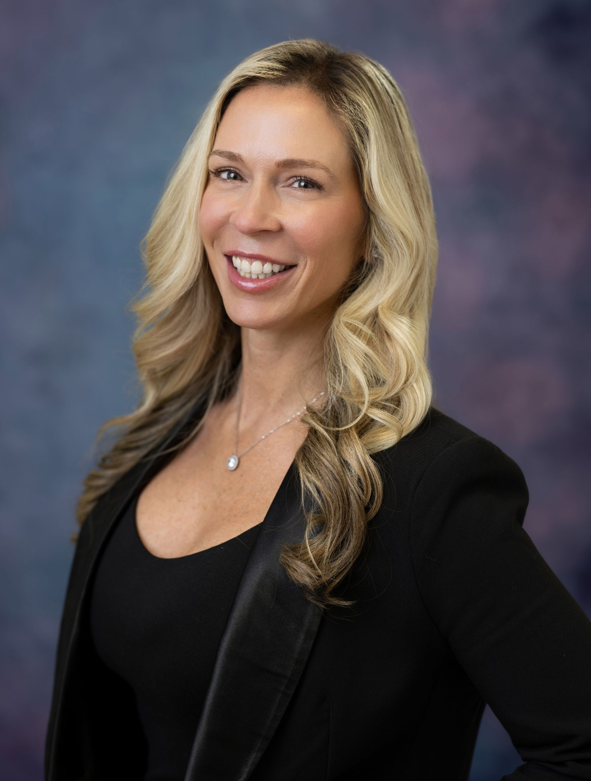 A woman with long blonde hair smiles, wearing a patterned sweater, against a blue background.