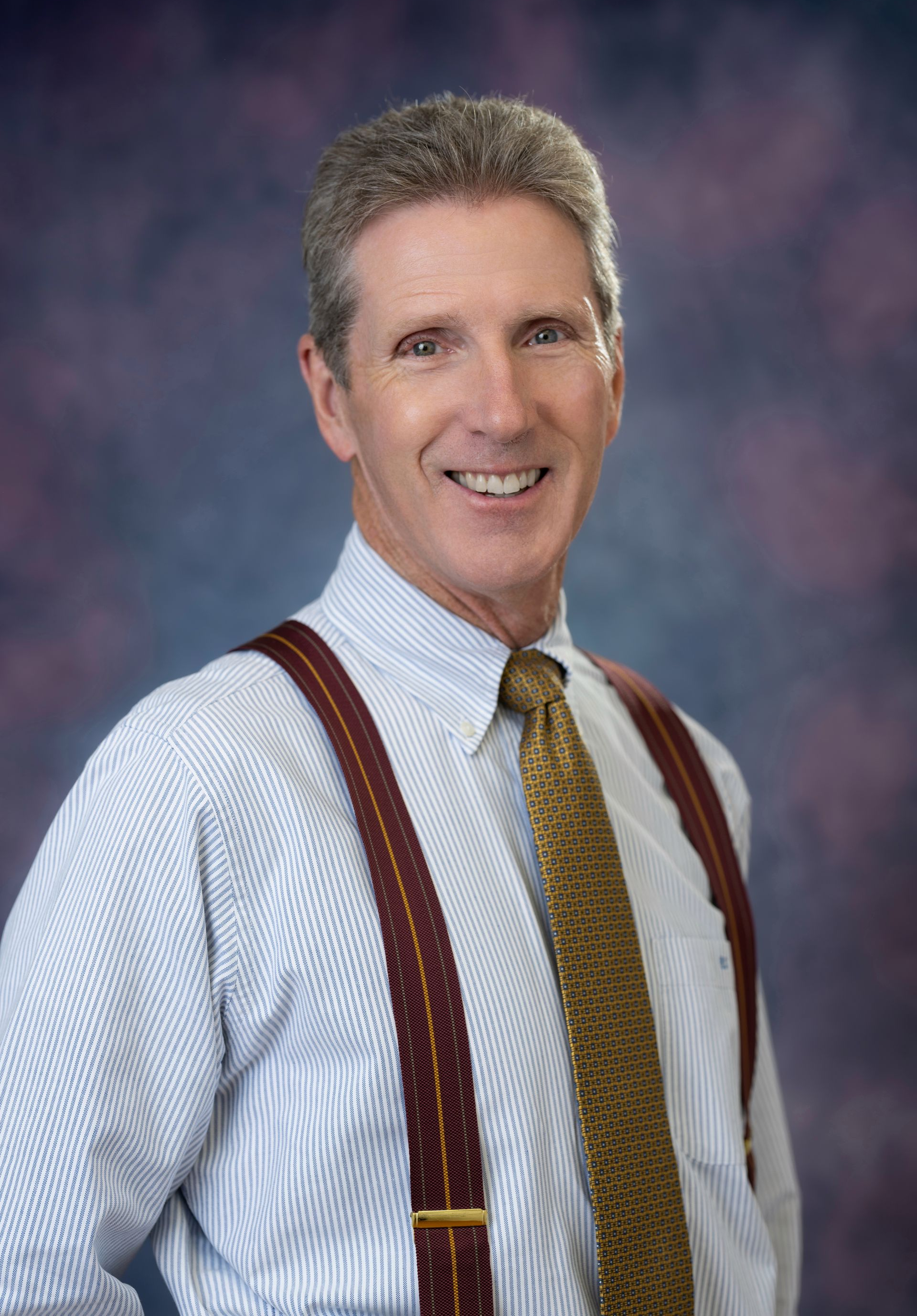 A smiling man with graying hair wearing a white shirt, patterned tie, and suspenders, against a blurred background.