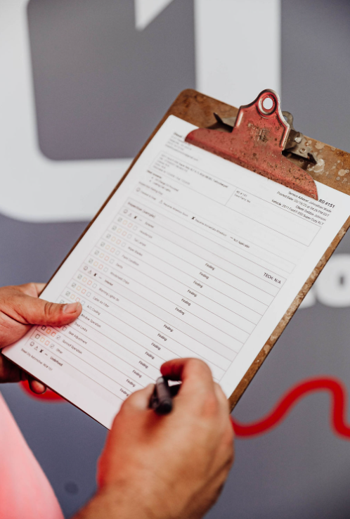 A person is writing on a clipboard in front of a car engine.