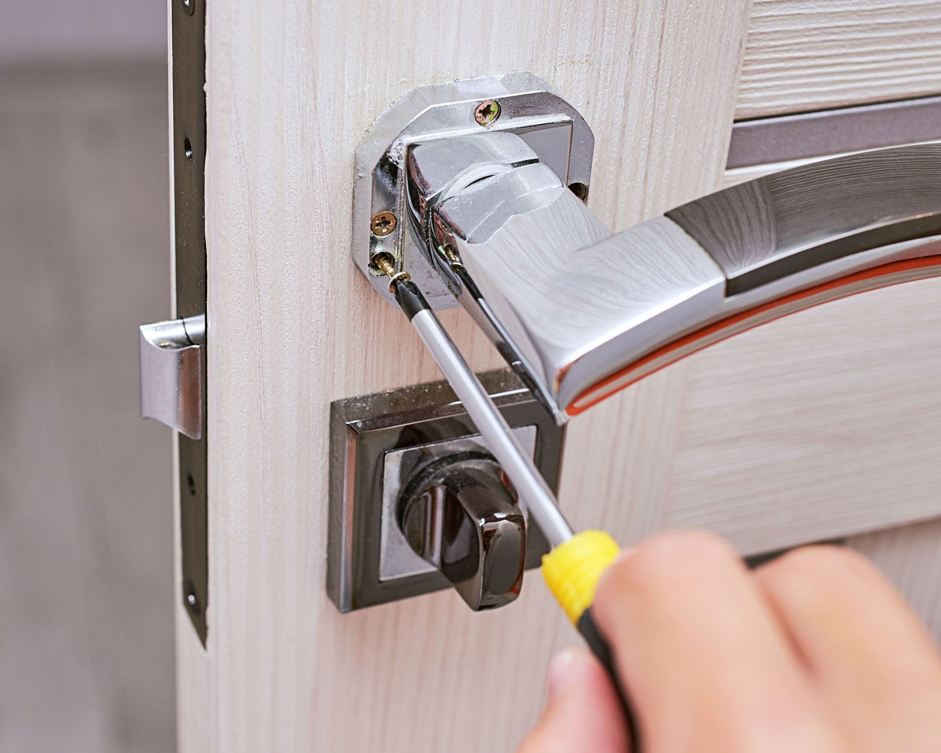 Person using a screwdriver to remove screws from a silver door handle on a light wood door.