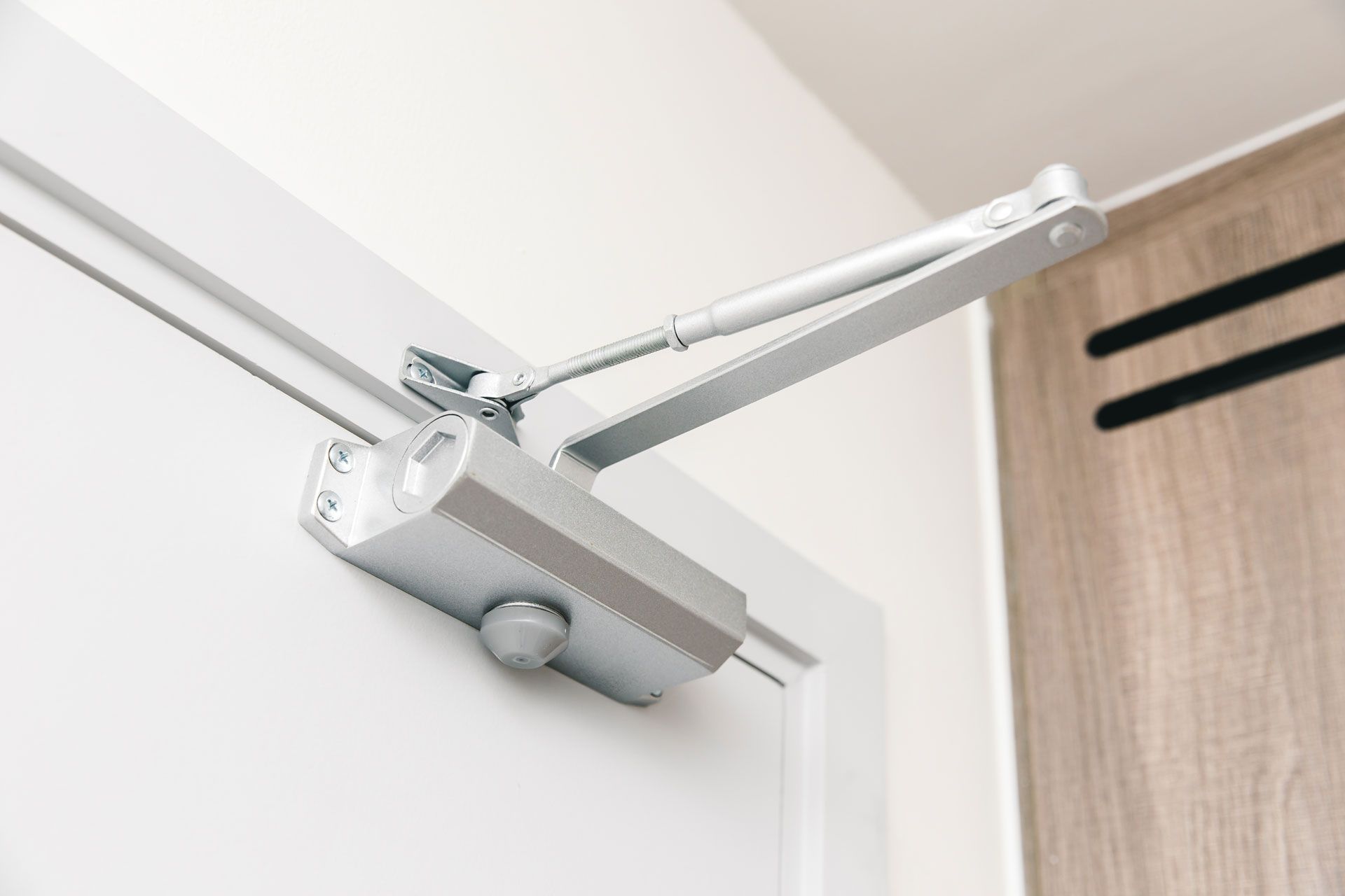 Silver door closer mounted on a white door frame, connected to a wooden door.