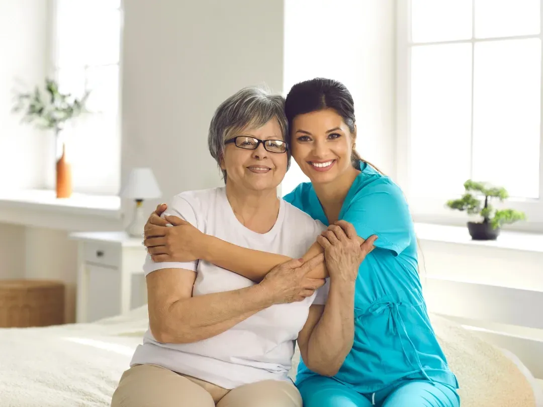 Two women embracing on a bed in a bright bedroom, smiling at the camera.