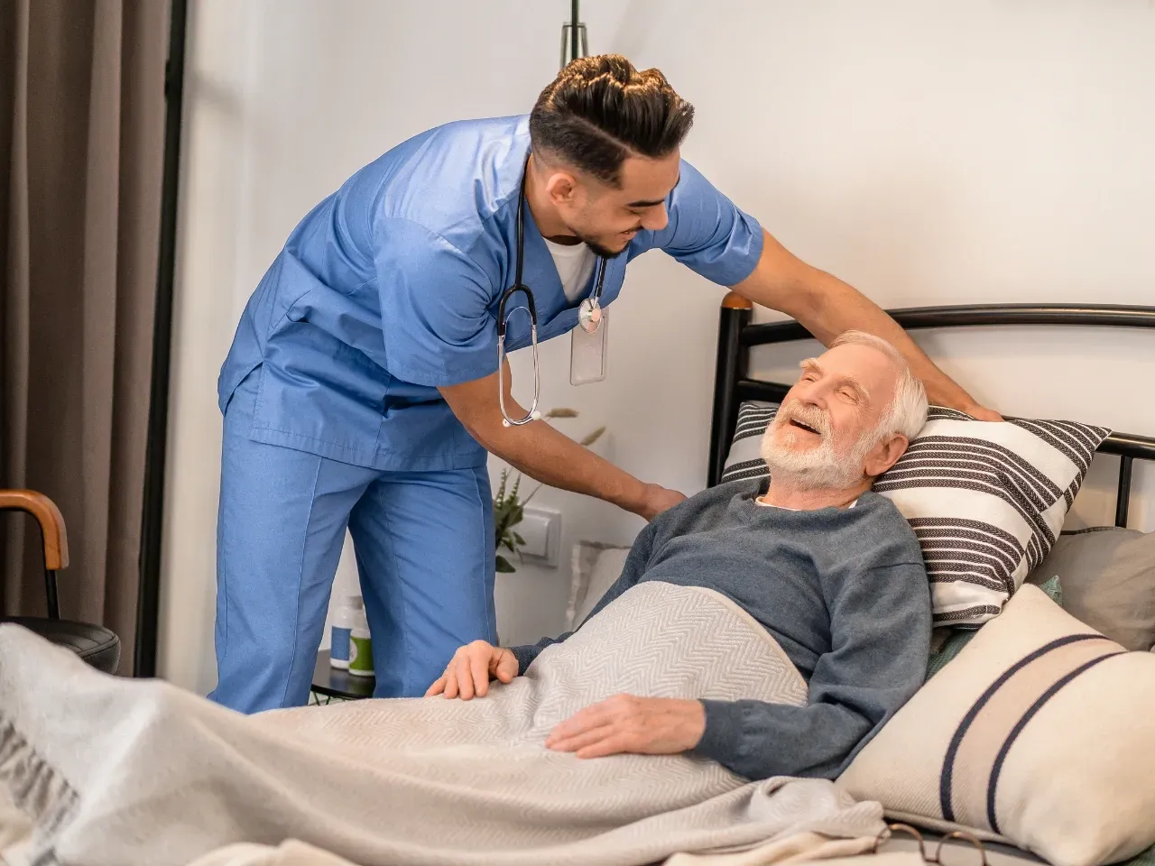 Nurse in blue scrubs checking an elderly man’s temperature in a hospital bed