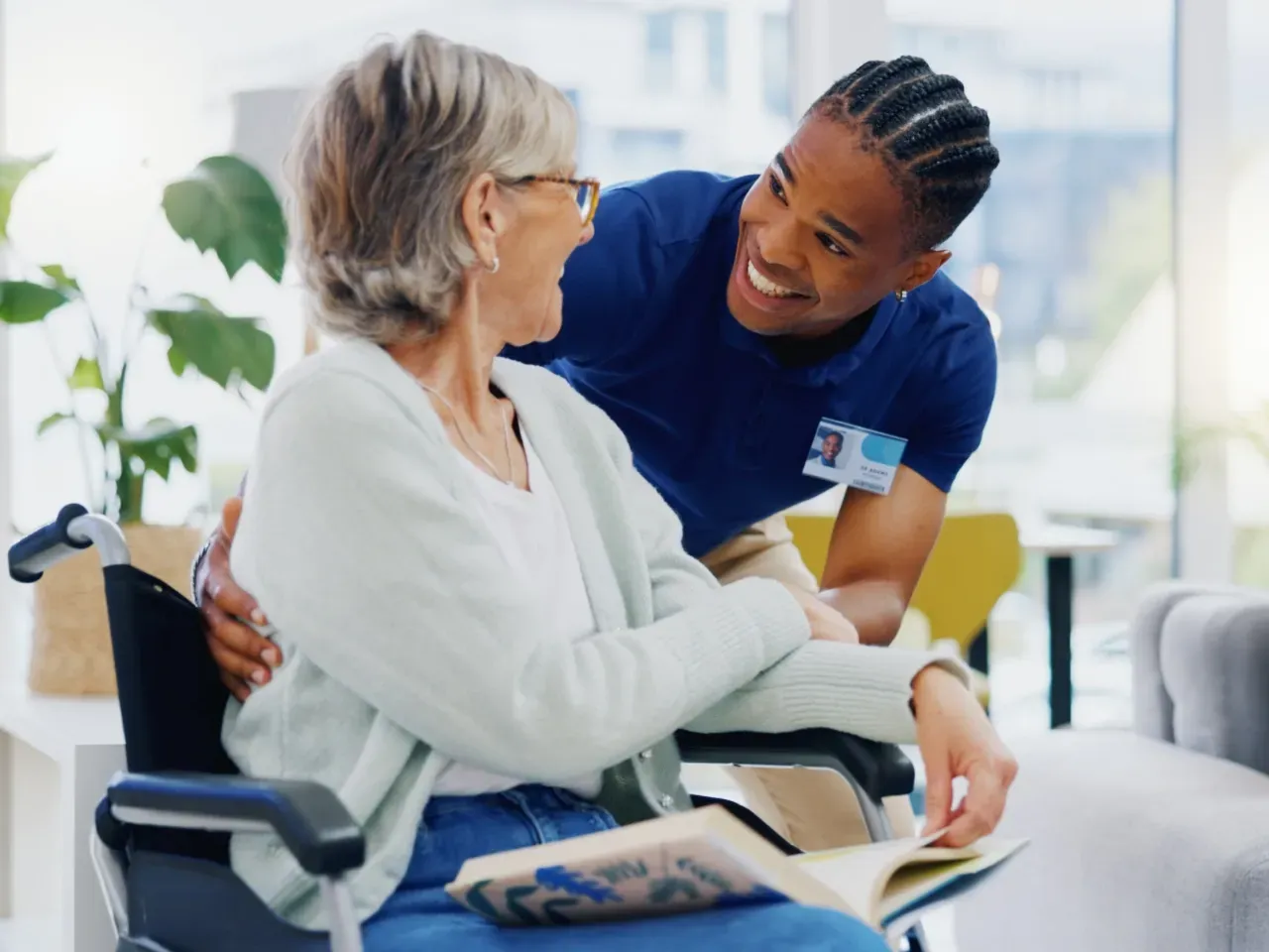 Nurse smiling and talking with a seated patient in a bright clinic, holding her hand and a tablet on her lap