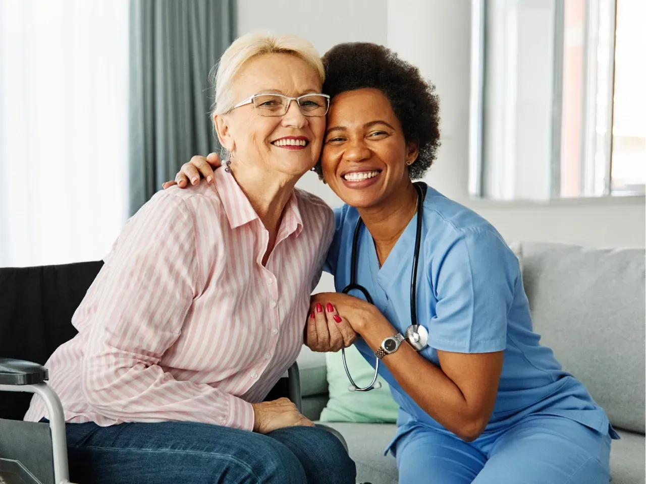 Two smiling healthcare workers in scrubs sitting together on a bed and holding hands
