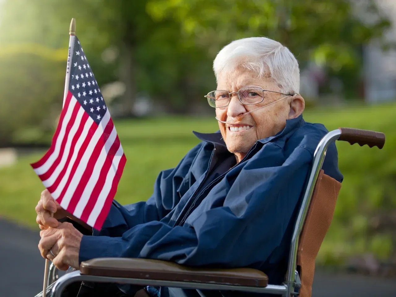 Elderly woman seated outdoors, smiling and holding a small American flag.