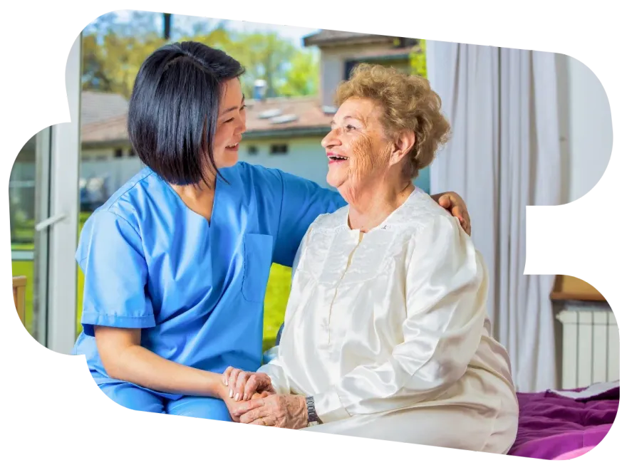 Nurse in blue scrubs holding hands with an elderly woman in white, smiling in a hospital room