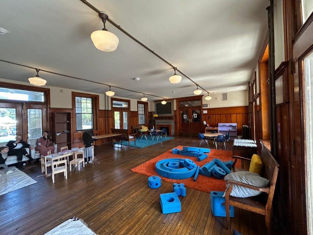 Bright indoor playroom with blue foam play set, tables by windows, and wooden floors