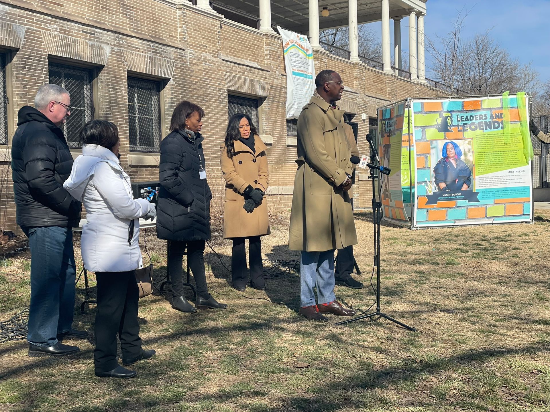 People gathered outdoors beside a memorial poster on a lawn in front of a building.