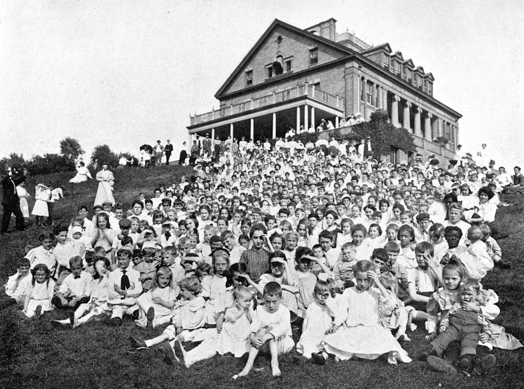 Large group posed on a hillside in front of a large house with a porch.