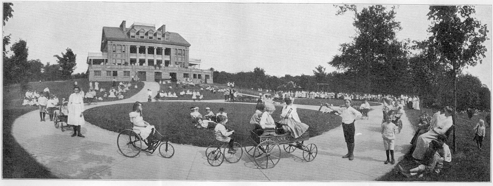 Cyclists and pedestrians on a curved road beside a large building and trees in a vintage black-and-white scene