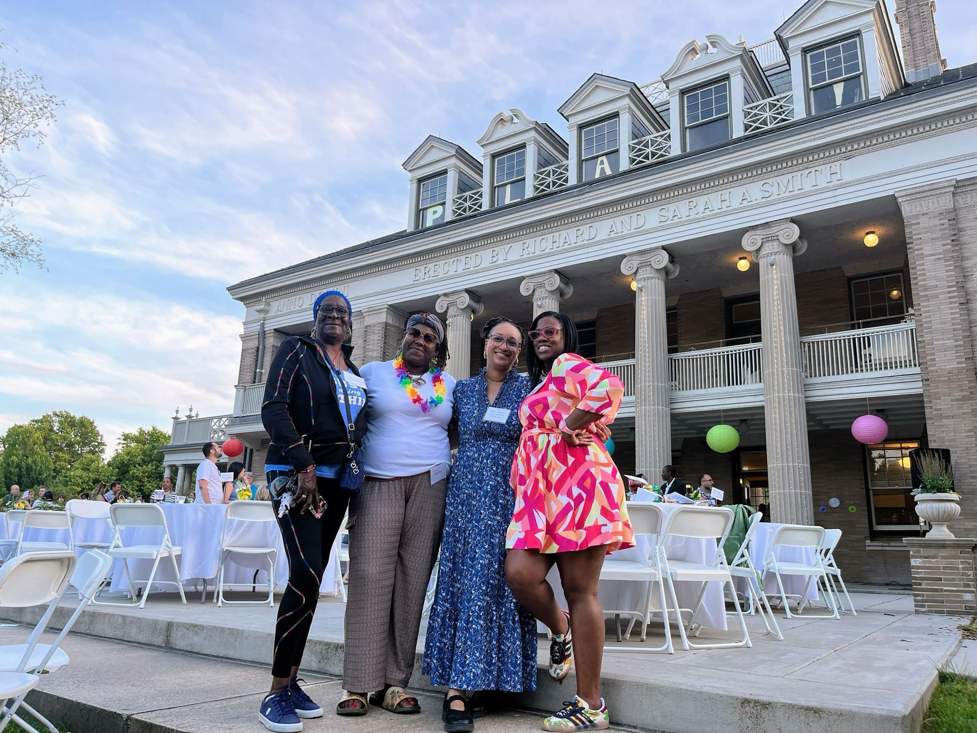 Four people posing on steps outside a grand building with white tables and chairs on the patio.