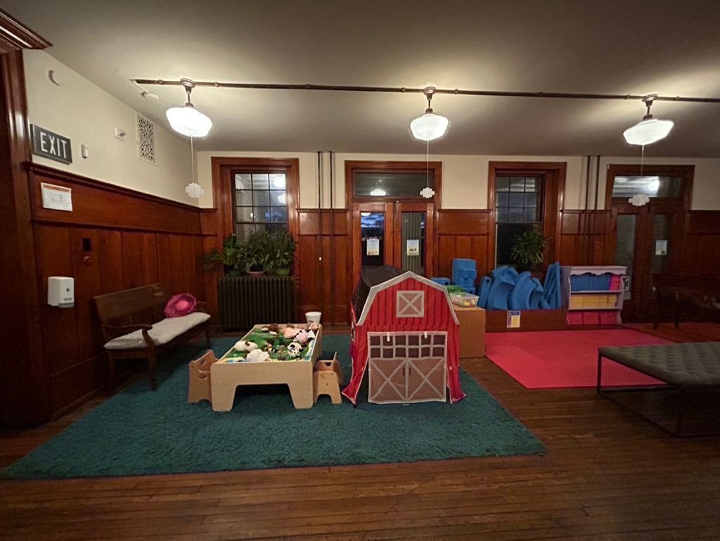 Indoor playroom with toy furniture, a red playhouse, and colorful mats under ceiling lights