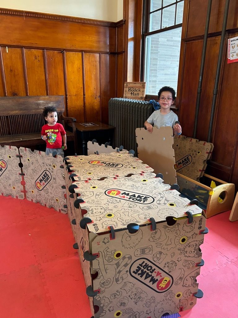 Two children in a wooden indoor play area behind a cardboard obstacle course