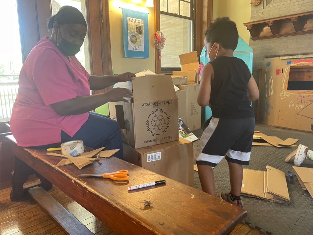 Two people assembling a cardboard box at a table in a room with craft supplies.