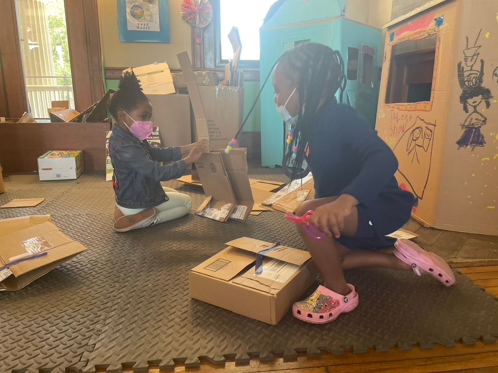 Two children kneel on a carpet, opening cardboard boxes in a classroom-like room with shelves and artwork.