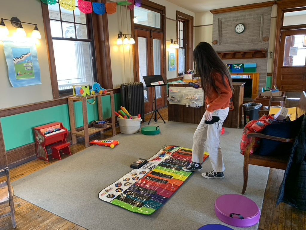 Person standing on a colorful play mat in a cozy room with windows, chairs, and toys.